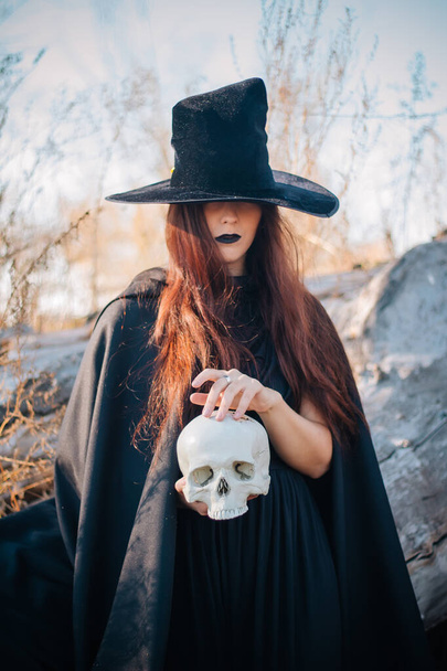 A young witch with pale skin and black lips in a black hat, a dress and a raincoat. Holds the skull of a dead man. Autumn, fallen tree and tall, dry grass. Halloween, magic, fantasy. - Photo, Image