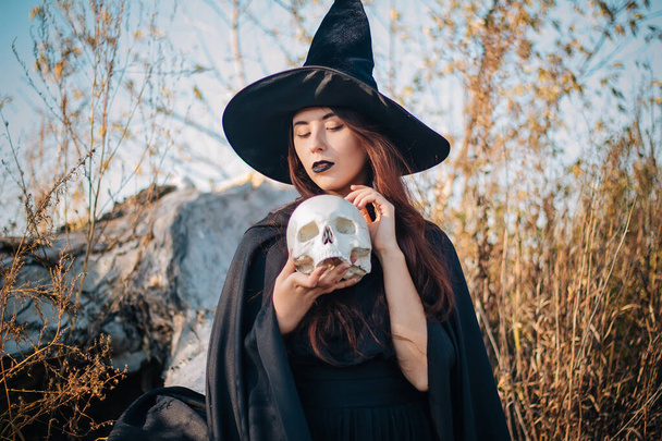 A young witch with pale skin and black lips in a black hat, a dress and a raincoat. Holds the skull of a dead man. Autumn, fallen tree and tall, dry grass. Halloween, magic, fantasy. - Foto, afbeelding