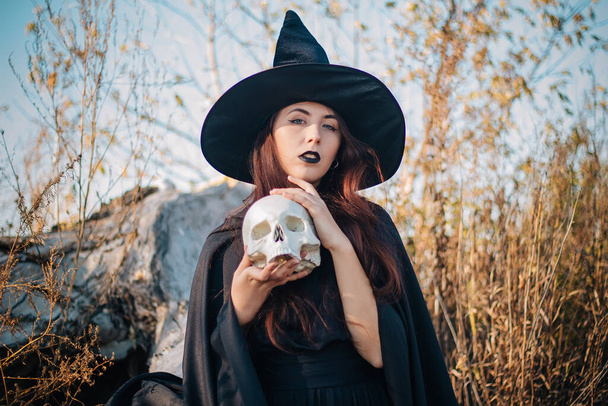 A young witch with pale skin and black lips in a black hat, a dress and a raincoat. Holds the skull of a dead man. Autumn, fallen tree and tall, dry grass. Halloween, magic, fantasy. - Фото, зображення