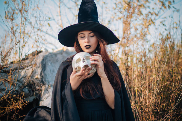 A young witch with pale skin and black lips in a black hat, a dress and a raincoat. Holds the skull of a dead man. Autumn, fallen tree and tall, dry grass. Halloween, magic, fantasy. - Foto, immagini