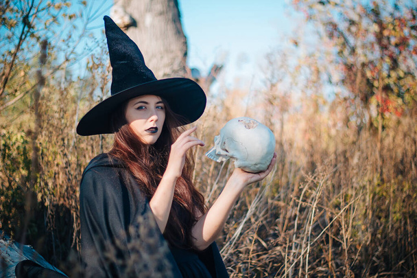A young witch with pale skin and black lips in a black hat, a dress and a raincoat. Holds the skull of a dead man. Autumn, fallen tree and tall, dry grass. Halloween, magic, fantasy. - Photo, image