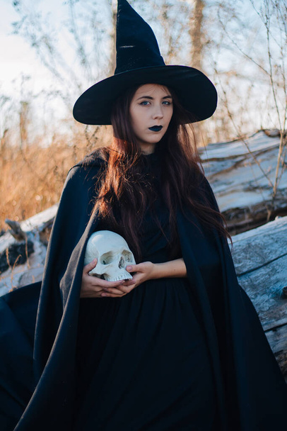 A young witch with pale skin and black lips in a black hat, a dress and a raincoat. Holds the skull of a dead man. Autumn, fallen tree and tall, dry grass. Halloween, magic, fantasy. - Photo, Image