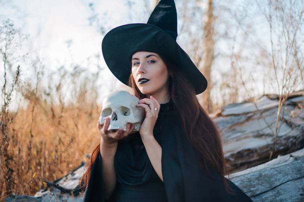 A young witch with pale skin and black lips in a black hat, a dress and a raincoat. Holds the skull of a dead man. Autumn, fallen tree and tall, dry grass. Halloween, magic, fantasy. - Fotó, kép