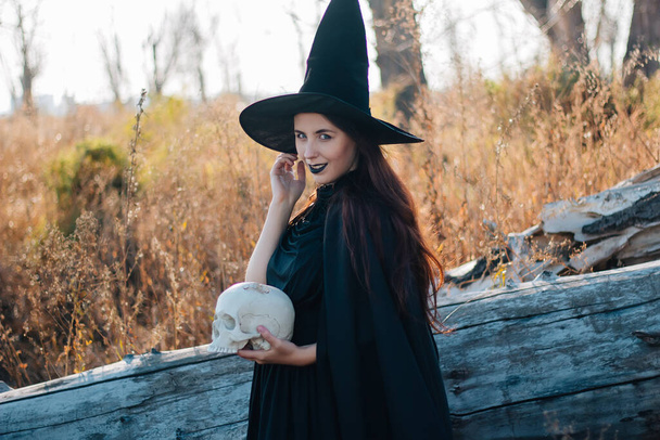 A young witch with pale skin and black lips in a black hat, a dress and a raincoat. Holds the skull of a dead man. Autumn, fallen tree and tall, dry grass. Halloween, magic, fantasy. - Zdjęcie, obraz