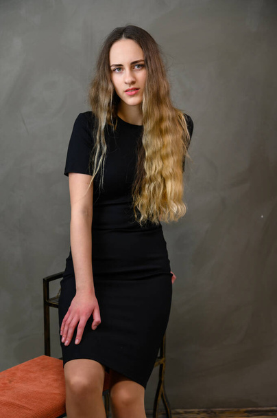 Studio photo of a caucasian young pretty blonde girl in a black dress with long hair sitting on a stand against a gray background. - Fotó, kép