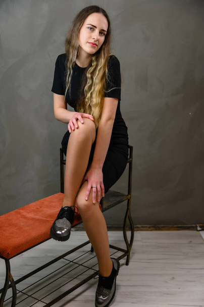 Studio photo of a caucasian young pretty blonde girl in a black dress with long hair sitting on a stand against a gray background. - Foto, Imagem