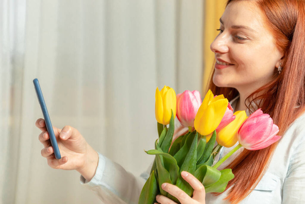 Womens Day March 8. Valentines day, birthday concept. Young beautiful cute long hair girl holding a fresh bouquet of tulips and taking selfie photo, smiling at camera - Foto, Imagen