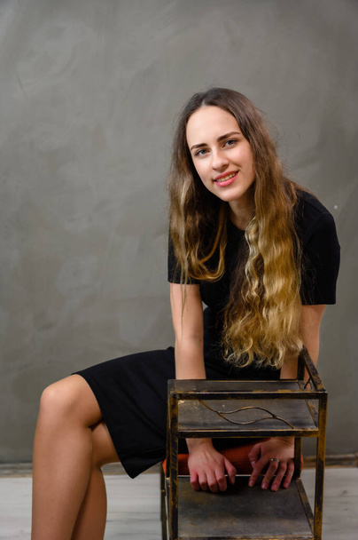 Vertical portrait of a caucasian girl in a black dress with long hair sitting on a stand on a gray background. - Фото, изображение