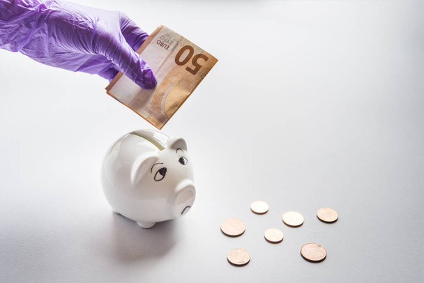 Hand in glowes put money to sad piggy bank - Foto, Bild