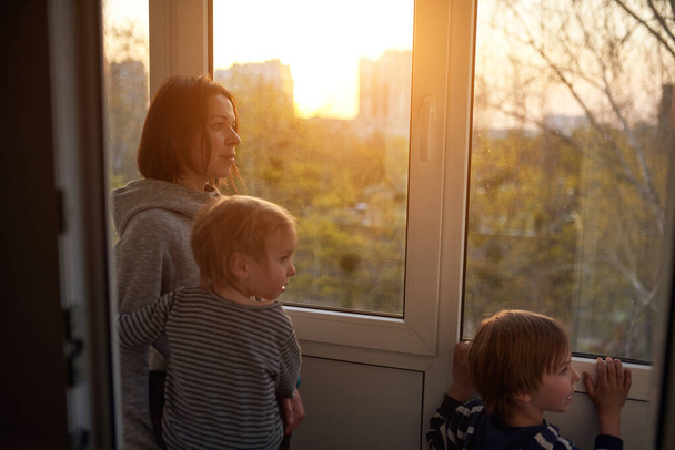 Mother with two sons looks out the window at sunset in isolation at home for virus outbreak. Stay home concept. - Fotoğraf, Görsel