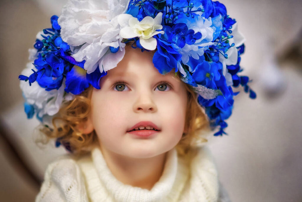 Κοντινό πλάνο πορτραίτο μικρού 4χρονου ξανθού σγουρού κοριτσιού με ζεστό πουλόβερ και μεγάλο τεχνητό στεφάνι με μπλε και λευκά λουλούδια. Περιμένω την άνοιξη. Χειμερινό πορτρέτο. Όμορφο παιδί με μεγάλα μάτια. - Φωτογραφία, εικόνα