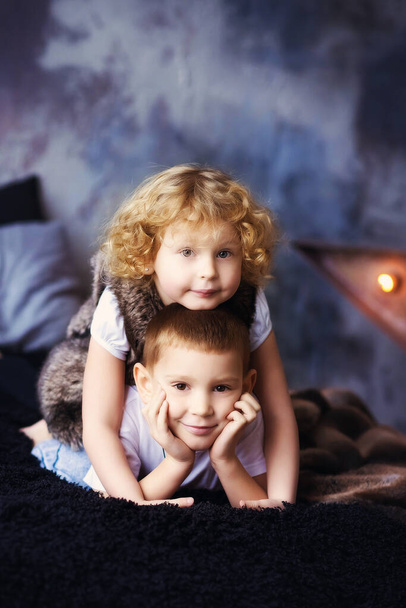 Vertical portrait of two children, boy and girl, brother and sister, siblings, laying in bed in loft styled bedroom. Curly blonde girl and her brother. Kids spend time together. Family. Self isolation - Valokuva, kuva