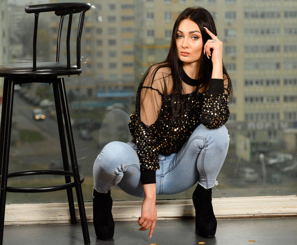 Photo model glamorous girl in brunette jeans posing on a chair against the background of a window - Fotoğraf, Görsel