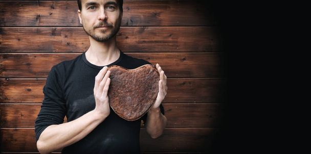 αρτοποιός κρατώντας στα χέρια vegan σίκαλη ολικής αλέσεως χωρίς μαγιά ψωμί προζύμι σε σχήμα καρδιάς κοντά στο στήθος του, σε φόντο από καφέ ξύλινο τοίχο, χειροποίητο ιδιωτικό φούρνο, δώρο ημέρα του Αγίου Βαλεντίνου, αντίγραφο χώρο - Φωτογραφία, εικόνα