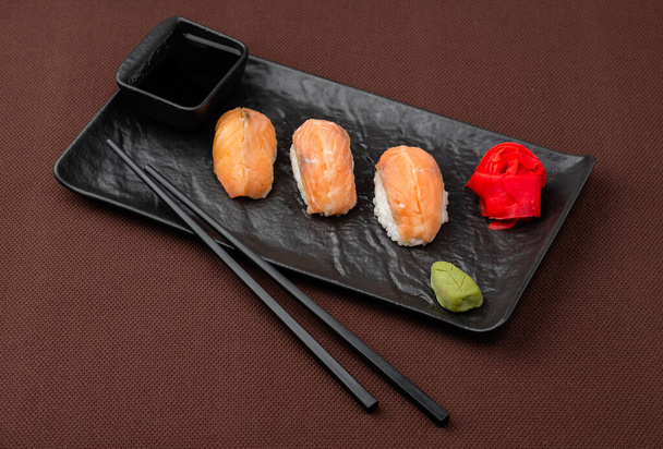 sushi sashimi con salmón, jengibre, wasabi, salsa de soja y palillos para sushi en un plato negro sobre un fondo textil marrón
 - Foto, imagen