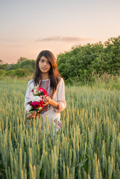 Smiling indian girl wearing traditional Ukrainian embroidery dress clothes posing in field, beautiful ethnic young woman in nature hold handmade flower wreath or bouquet in hands - Photo, Image