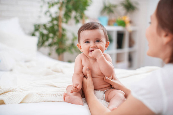 Cute smiling baby with scratching teeth on the bed in the room. The mother supports the child while sitting. The baby is just starting to sit on its own.The concept of support and care. - Valokuva, kuva