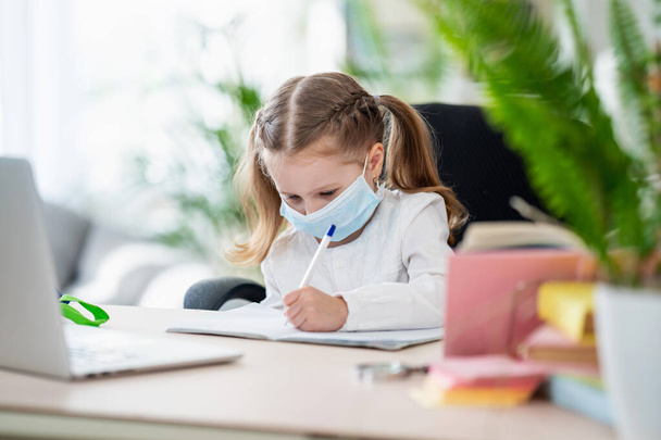 Cute little girl, doing homework, writing in a notebook , using a laptop, digital e -learning concepts, during quarantine and self- isolation. Remote e-learning due to Covid-19 coronavirus pandemic - Foto, Imagem