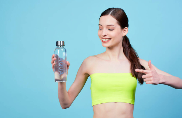 Image of cute attractive athletic young Caucasian girl in a light green top, holding a water bottle in her right hand and pointing to it with the finger of her left hand, posing on a blue background. - Foto, afbeelding
