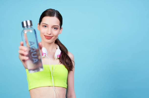 Charming athletic young Caucasian girl, in headphones and a light green top, posing on a blue background, holding a water bottle with the word aqua. Copy space. - Foto, Imagen