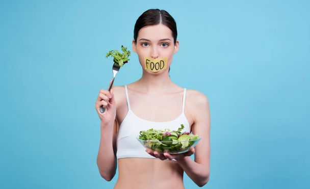 Sad pretty young girl in a white top, holding a bowl with a fresh green salad and a fork in her hand, her mouth is sealed with a ribbon with the word food, she can t eat, posing on a blue background. - Photo, Image