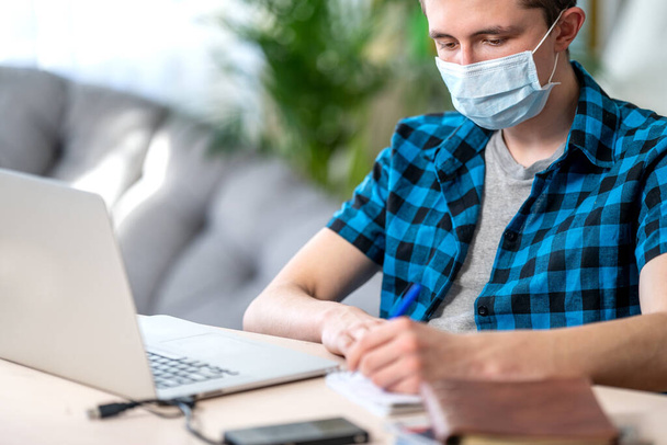 active teenager in a mask with homework on a laptop during a coronavirus quarantine. Social distance, self-isolation. Distance learning due to virus, COVID-19 - Photo, image
