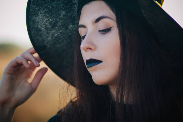 Porträt eines schönen Mädchens mit blasser Haut und schwarzen Lippen in einem großen schwarzen Hut. Frau als Hexe zu Halloween. - Foto, Bild