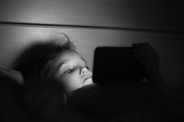 a little girl watches a cartoon or educational program before going to bed, phone and gadgets, bed and bed, the impact of the new on the child - 写真・画像
