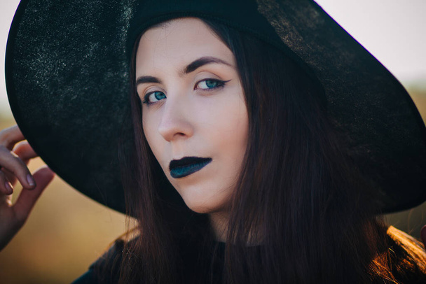 Portrait of a beautiful girl with pale skin and black lips in a big black hat. Woman in the image of a witch for Halloween. - Foto, Bild