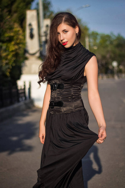 Young, beautiful woman in a black dress with a corset, walking along a city street against a background of a park and stone columns with lanterns. - Фото, зображення