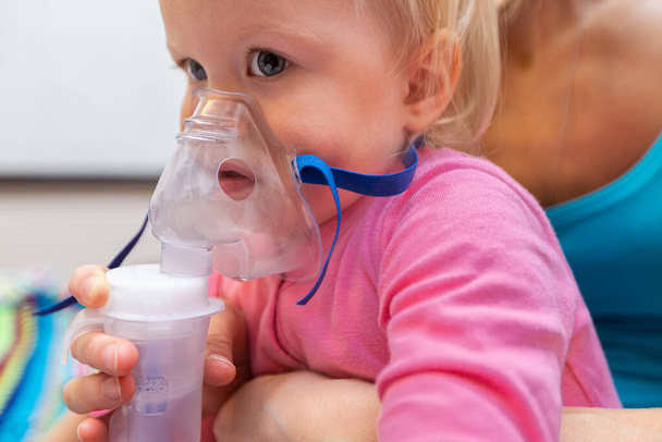 Two year old girl making inhalation with nebulizer at home with mother. The treatment of child with inhaler and nebulizer steam sick from cough and sore throat. - Photo, Image