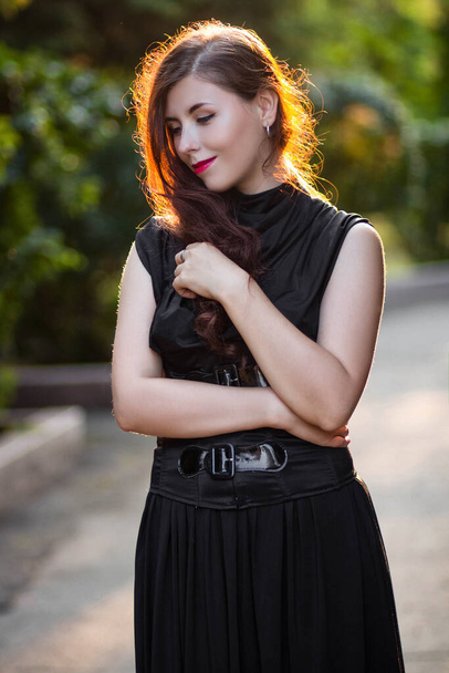 Beautiful, young woman in a black dress with a corset walking in the park in the evening at sunset. - Photo, Image