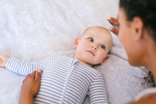 Portrait of toddler lying on bed with her mother. Close up face of cute relaxed infant looking at mother playing with copy space. Rear view of multiethnic woman playing with adopted child lying on bed. - Foto, Imagem