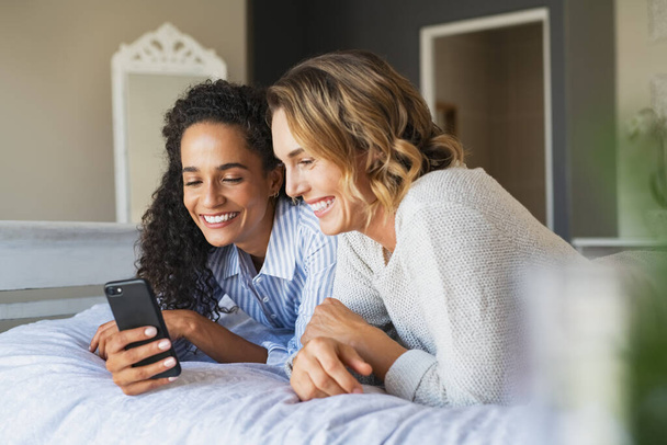 Cheerful multiethnic female friends doing online shopping while lying on bed and using smartphone. Two young women watching funny videos on mobile phone and laughing together. Lesbian couple using cellphone while relaxing on bed. - Foto, immagini