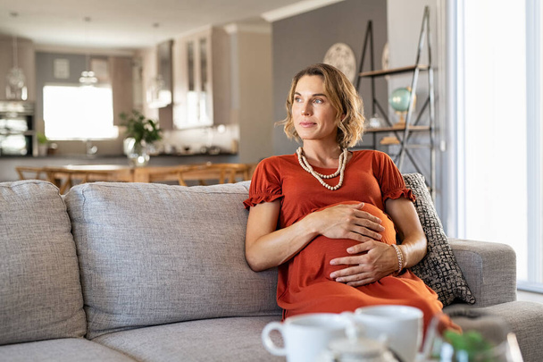 Thoughtful middle aged pregnant woman sitting on couch looking away with copy space. Pensive expecting mother relaxing on sofa at home. Beautiful pregnant woman with hands on baby bump thinking about unborn child. - Foto, imagen