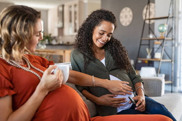Smiling woman with hands on expecting mother's stomach feeling baby movement while sitting on couch at home. Lesbian couple both with a baby bump relaxing at home. Happy friend feeling belly of young african pregnant woman.  - Photo, image