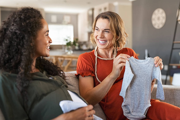 Happy pregnant woman looking at baby clothes with joyful african friend. Laughing pregnant friends sitting on sofa and looking at baby shower gifts. Excited two expecting girls checking baby clothes for new baby.  - Foto, immagini