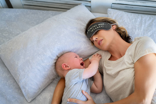 Tired mother with eye band trying to sleep while little toddler crying on bed. Angry baby boy crying and trying to wake mother. Stressed mother trying to take a nap with her child. - Fotó, kép