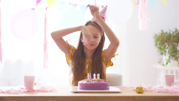 Blank meisje lacht dromerig en kijkt naar verjaardagsregenboogtaart. Feestelijke kleurrijke achtergrond met ballonnen. Verjaardagsfeest en wensenconcept. - Video