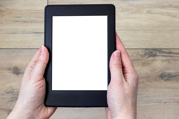 A modern black electronic book with a white blank screen in female hands against a blurred wooden tile floor background. Mockup tablet closeup - Photo, Image