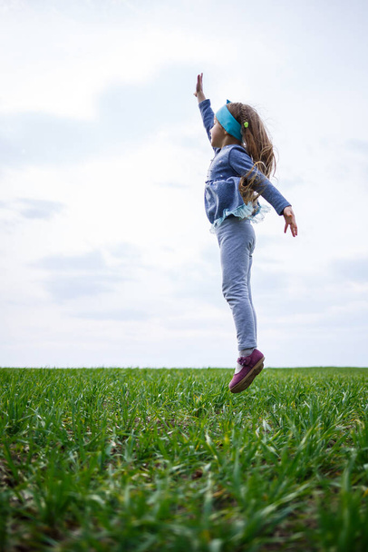 niña pequeña corre y salta, hierba verde en el campo, clima soleado de primavera, sonrisa y alegría del niño, cielo azul con nubes
 - Foto, Imagen