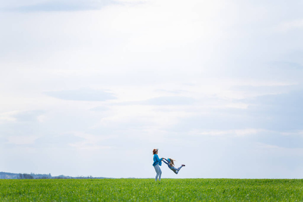 κοριτσάκι παιδί και μητέρα γυναίκα τρέχει και άλμα, πράσινο γρασίδι στο πεδίο, ηλιόλουστη άνοιξη καιρός, χαμόγελο και χαρά του παιδιού, μπλε ουρανό με σύννεφα - Φωτογραφία, εικόνα