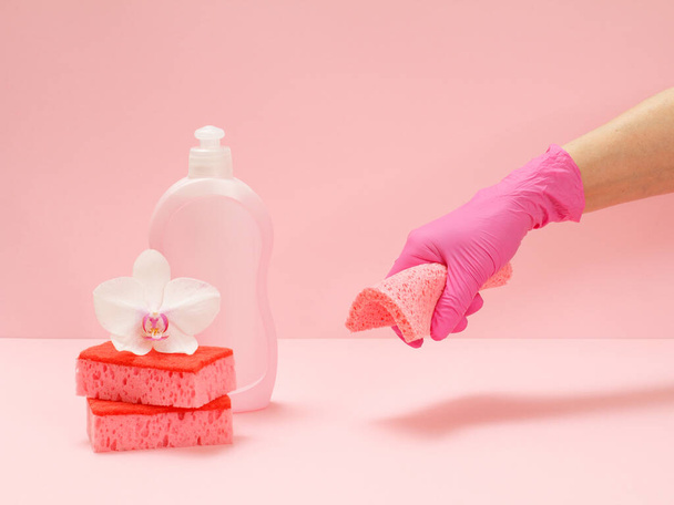 Botella de plástico de líquido lavavajillas, esponjas rojas, una flor de orquídea blanca y una mano en un guante de goma sosteniendo un trapo sobre el fondo rosa. Juego de lavado y limpieza
. - Foto, imagen