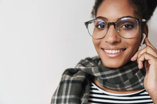 Portrait closeup of african american joyful woman in eyeglasses smiling and using earpod isolated over white background - Fotó, kép