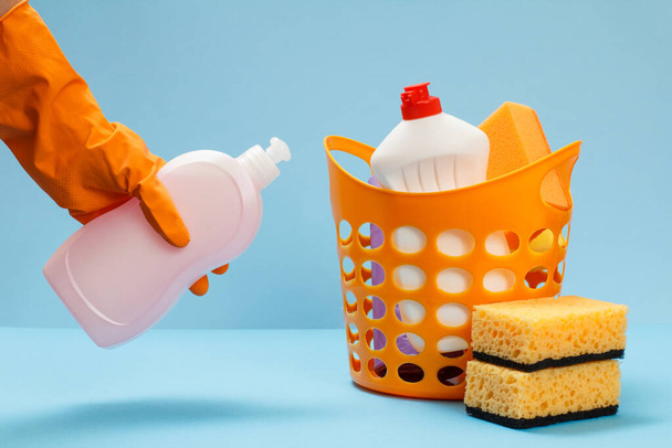Main de femme en gant de protection orange tenant une bouteille de liquide vaisselle avec un ensemble de lavage et de nettoyage sur le fond bleu
. - Photo, image
