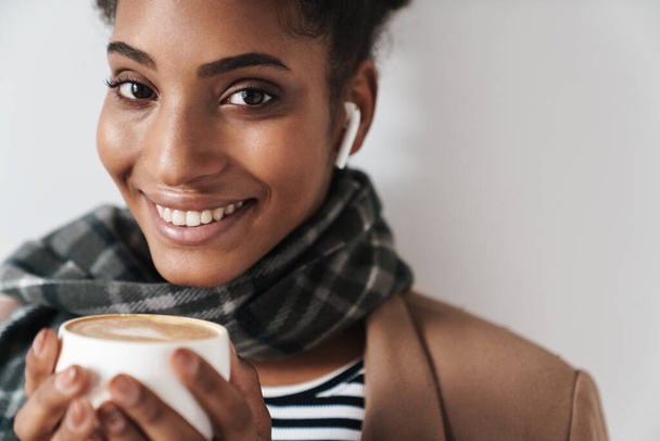 Portrait of african american smiling woman drinking coffee and using earpod isolated over white background - Zdjęcie, obraz