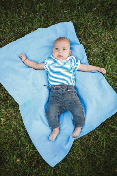 Ένα αγοράκι τριών μηνών ξαπλώνει ανάσκελα σε ένα μπλε κάλυμμα κρεβατιού στο πάρκο γύρω από το πράσινο γρασίδι και τα δέντρα. Παιδικά συναισθήματα χαράς - Φωτογραφία, εικόνα