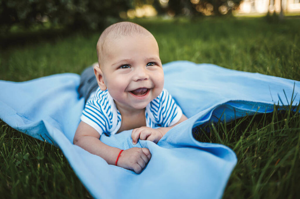 Niño de 3 meses de edad yace boca abajo sobre una colcha azul en el parque alrededor de la hierba verde y los árboles. Las emociones de alegría de los niños
 - Foto, Imagen