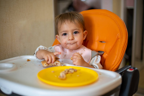 Χαριτωμένο μωρό σε μια πορτοκαλί καρέκλα μωρό μασάει εντατικά ένα κομμάτι κέικ με βρώμικο πρόσωπο. ψίχουλα και ένα πορτοκαλί πιάτο με μια πίτα στο τραπέζι. κοντινό πλάνο, μπροστινή όψη, μαλακή εστίαση, θολό φόντο - Φωτογραφία, εικόνα