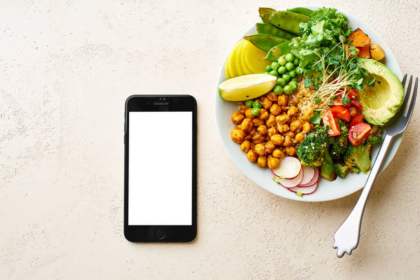 Flat lay with black smartphone and healthy vegetarian lunch bowl with avocado and chickpeas with quinoa and vegetables garnished with microgreens and nut dressing. Nutrient food delivery concept  - Фото, изображение